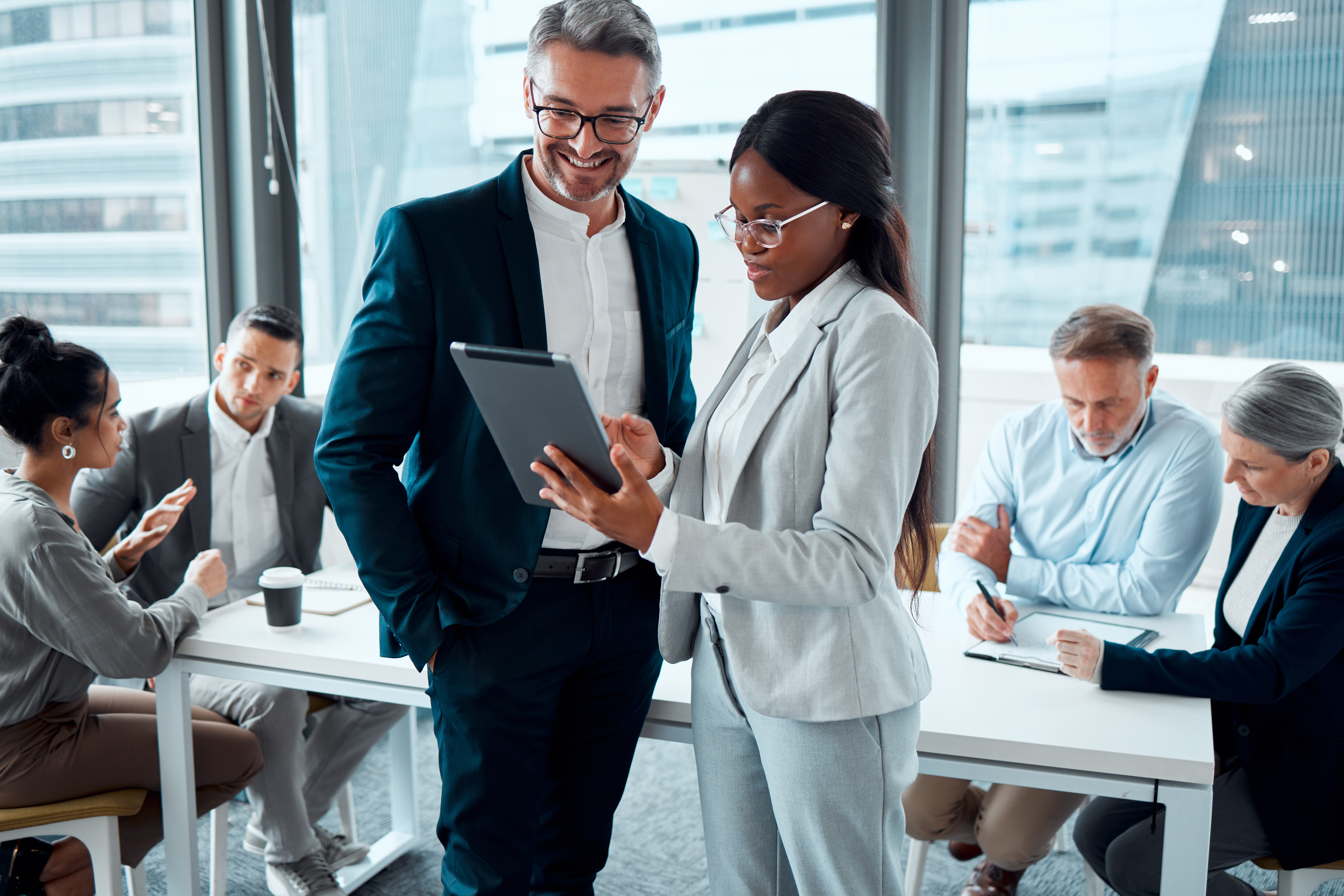 A woman dressed in a blazer standing next to a smiling man. She is showing him a digital audit management system easily with a tablet