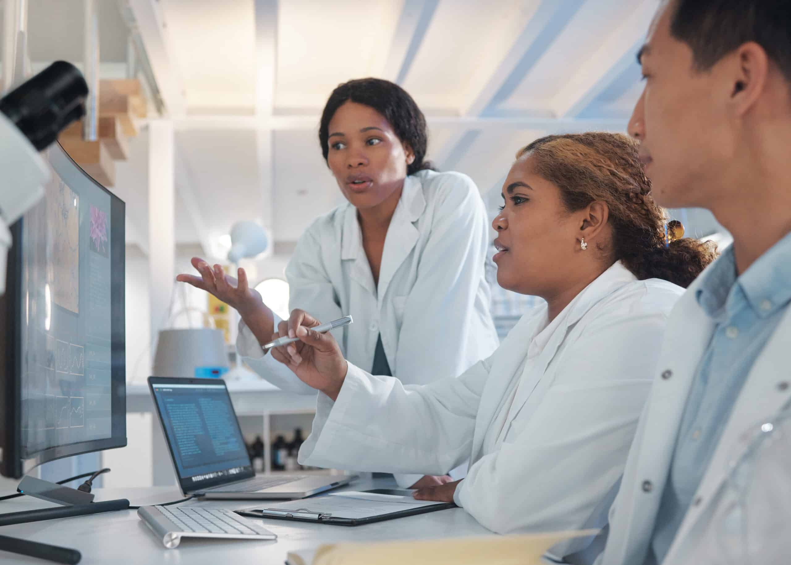 Three life sciences workers in lab coats pointing at a computer screen to discuss a complaint