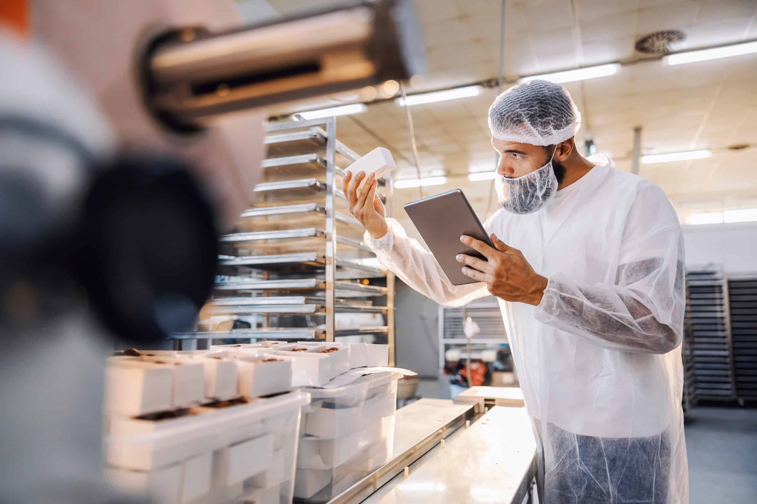A man with hair nets checking on the quality of a product while holding a tablet to log any deviations