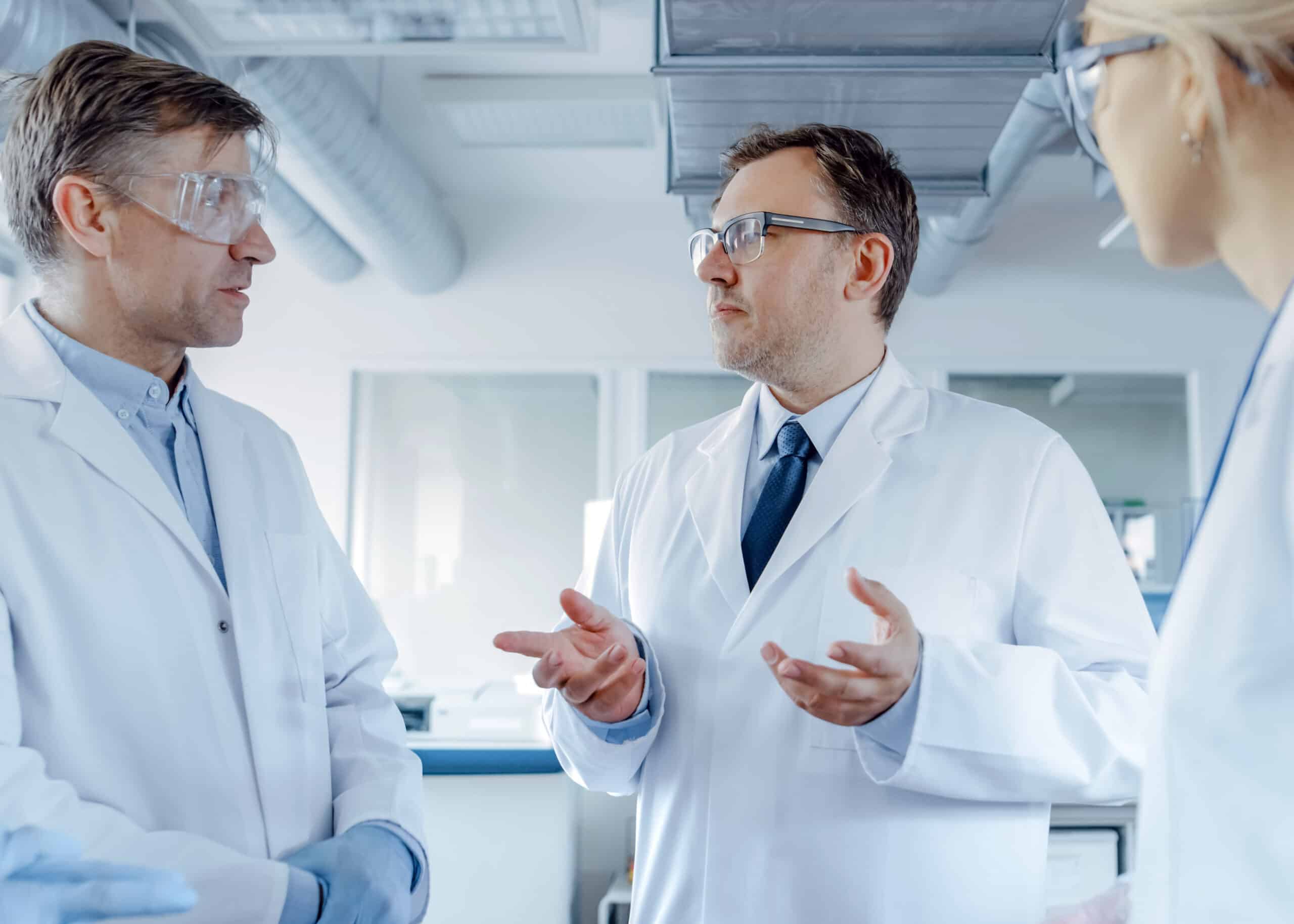 Three workers in lab coats and protective eye wear discussing nonconformances with focused expressions clear communication